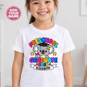 May include: A white t-shirt featuring a colourful graphic of a koala wearing a graduation cap and holding a diploma. The text reads "PRESCHOOL GRADUATE 2026" with a banner below displaying the name "ELIZABETH" and a badge saying "ADD YOUR NAME".