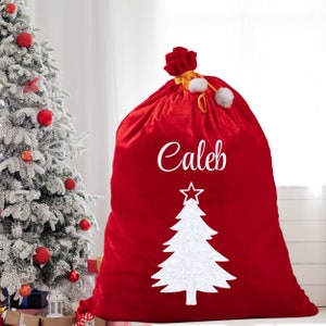 May include: A large red Christmas gift bag with the name "Caleb" in white script. A white Christmas tree design is below the name. The bag is tied at the top with a red bow and white pom-poms. A decorated Christmas tree is in the background.