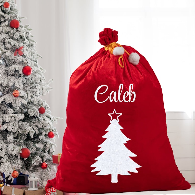 May include: A large red Christmas gift bag with the name "Caleb" in white script. A white Christmas tree design is below the name. The bag is tied at the top with a red bow and white pom-poms. A decorated Christmas tree is in the background.