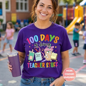 May include: A purple t-shirt with the text "100 DAYS of doing TEACHER STUFF" in colourful letters. The shirt features graphics of lesson plans, a coffee drink, and other teacher-related items. A purple tumbler is held in the hand.