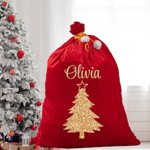 May include: A large red velvet Christmas gift bag with the name "Olivia" in gold script. A gold glitter Christmas tree design is centered on the bag. The bag is tied at the top with a gold cord and white pom-poms.