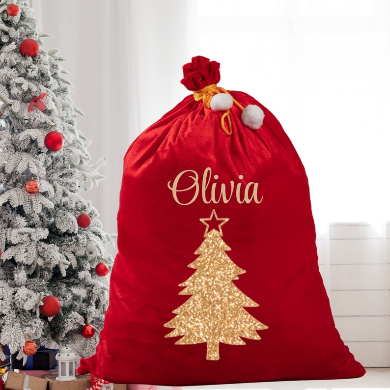 May include: A large red velvet Christmas gift bag with the name "Olivia" in gold script. A gold glitter Christmas tree design is centered on the bag. The bag is tied at the top with a gold cord and white pom-poms.