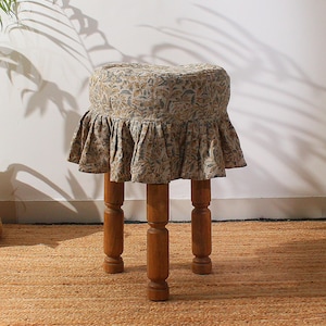 Op de afbeelding: Een kleine, ronde houten kruk met een bloemmotief zitting en een gerimpelde rok. Het zitkussen heeft een beige en bruin bloemenpatroon. De kruk heeft drie stevige, gedraaide houten poten. De kruk staat op een geweven vloerkleed.