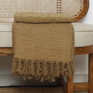May include: A folded, textured, tan throw blanket with fringe detail is draped over a bench. The bench has a light-coloured cushion and a wooden frame with a woven back. The blanket is made of a thick, woven material.