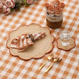 May include: A brown and white checkered tablecloth with a woven placemat, a napkin, and a glass of water. The placemat has a scalloped edge and is made of natural fibers. There is a wooden napkin ring on the napkin. The glass of water is on a smaller woven coaster. Two gold forks are on a wooden holder. A small basket with the word "Noah" on it holds rolled up napkins.