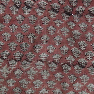 May include: A woven fabric featuring a repeating pattern of small, stylized floral motifs in a light beige color against a deep, earthy terracotta red background. The texture of the textile is visible, showing a consistent weave throughout the material.