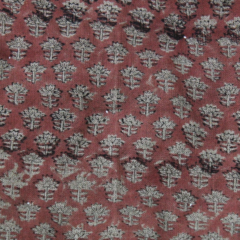 May include: A woven fabric featuring a repeating pattern of small, stylized floral motifs in a light beige color against a deep, earthy terracotta red background. The texture of the textile is visible, showing a consistent weave throughout the material.