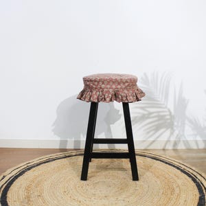 May include: A tall stool with four black legs and a round seat cushion. The cushion is covered in a dusty rose fabric featuring a repeating white floral pattern and is finished with a gathered, ruffled skirt that hangs around the seat edge.