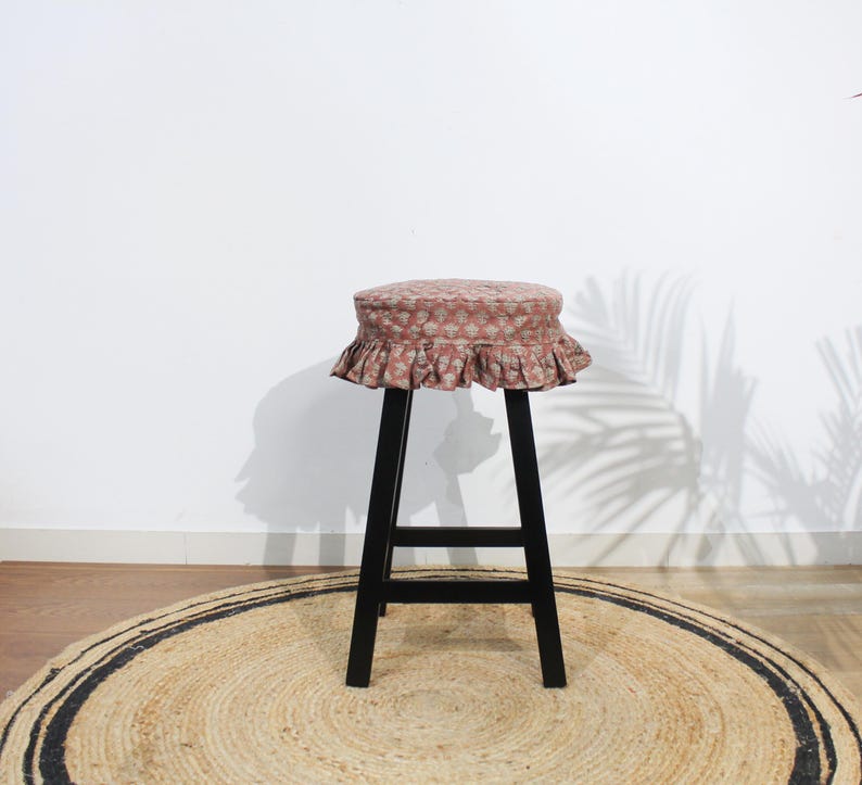 May include: A tall stool with four black legs and a round seat cushion. The cushion is covered in a dusty rose fabric featuring a repeating white floral pattern and is finished with a gathered, ruffled skirt that hangs around the seat edge.
