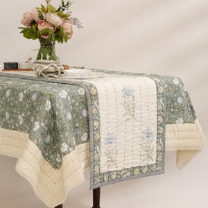 May include: A green and white floral tablecloth with a white quilted table runner featuring a floral design. The table runner has a light blue and yellow floral pattern. The tablecloth has a scalloped edge and a white trim.
