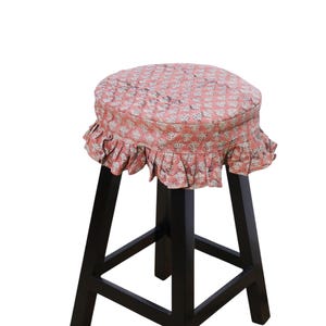 May include: A black wooden stool featuring a round seat cushion covered in a salmon-colored fabric with a repeating white floral block print pattern and a gathered, ruffled skirt that hangs around the edge of the circular seat top.