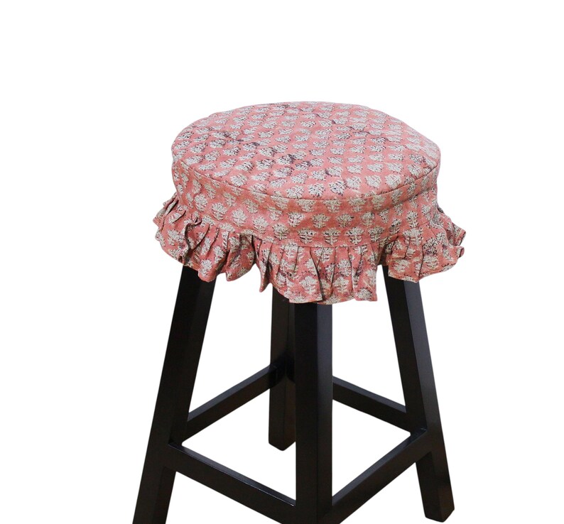 May include: A black wooden stool featuring a round seat cushion covered in a salmon-colored fabric with a repeating white floral block print pattern and a gathered, ruffled skirt that hangs around the edge of the circular seat top.