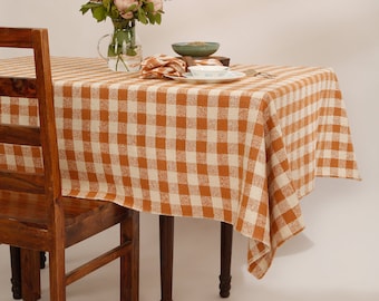 May include: A brown and white checkered tablecloth with a vase of pink flowers on a wooden table with two chairs.