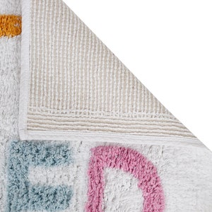 May include: A white bath mat with the word "BED" spelled out in blue, pink, and yellow. The mat has a non-slip backing.