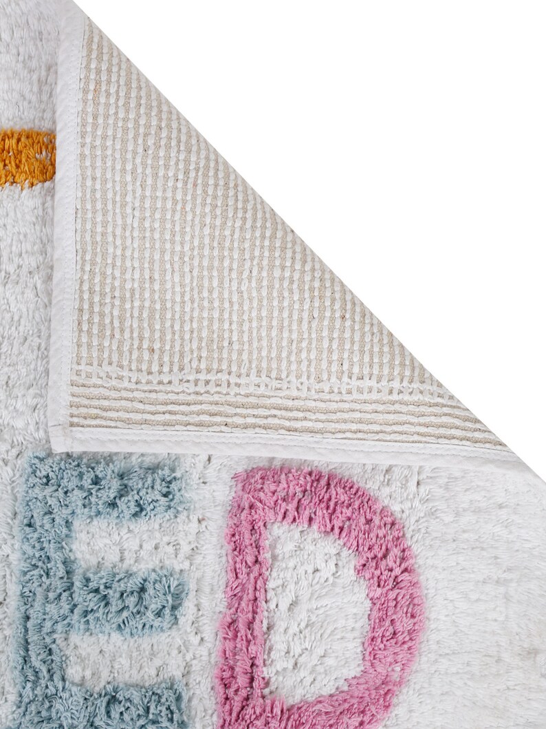 May include: A white bath mat with the word "BED" spelled out in blue, pink, and yellow. The mat has a non-slip backing.