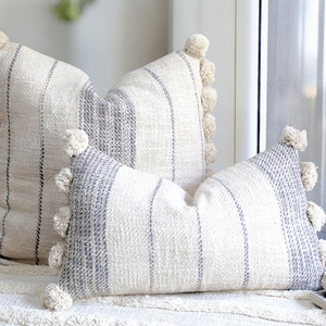 May include: Two striped cushion covers with pom-pom trim. The cushions are made of a soft, textured fabric and are a light cream colour with grey stripes.