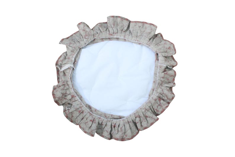 May include: A circular fabric cover featuring a white quilted center and a ruffled border with a mottled beige and reddish-brown pattern. The item has a gathered edge design and a soft, textured appearance, presenting a round shape overall.