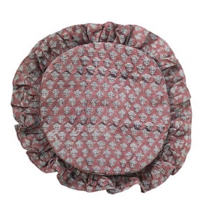 May include: A round, dusty rose-colored cushion featuring a repeating white floral block print pattern. The cushion is finished with a gathered, ruffled edge that encircles the entire perimeter, creating a soft, textured appearance.