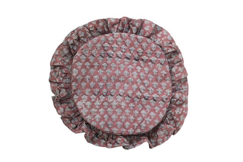 May include: A round, dusty rose-colored cushion featuring a repeating white floral block print pattern. The cushion is finished with a gathered, ruffled edge that encircles the entire perimeter, creating a soft, textured appearance.