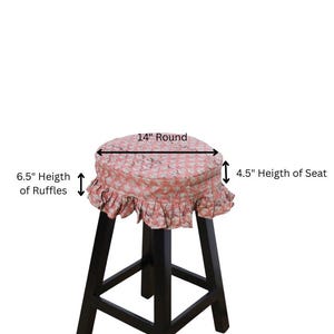 May include: A round stool cover with a pink and white floral pattern, featuring a 6.5-inch ruffle trim and a 4.5-inch seat height. The cover is 14 inches round and sits atop a black wooden stool with four legs and a square base support structure.