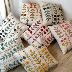 Puede incluir: Una colección de cojines decorativos con diseños florales bordados. Los cojines presentan tela color crema con bordados de varios colores, incluyendo verde, rojo, amarillo y azul. Algunos cojines tienen detalles de pompones.