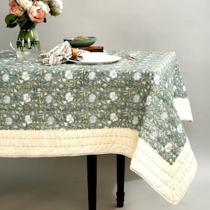 Könnte beinhalten: Eine grüne Tischdecke mit einem weißen Blumenmuster und einem cremefarbenen Rand. Die Tischdecke liegt auf einem Holztisch mit einer Blumenvase, einem Teller und einer Schüssel.