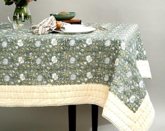 May include: A green tablecloth with a white floral pattern and a cream-colored border. The tablecloth is laid on a wooden table with a vase of flowers, a plate, and a bowl.