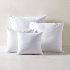 May include: Four white square pillows of different sizes, arranged in a pyramid shape on a white surface.