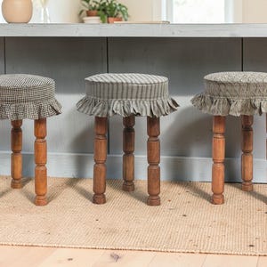 May include: Three wooden stools with patterned fabric seats and ruffled skirts. The seats feature different patterns: a black and white check, gray stripes, and a textured design. The stools have turned wooden legs.