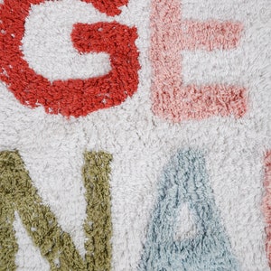 May include: A white bath rug with the word "GENA" spelled out in colorful, fluffy letters. The letters are red, pink, green, and blue.
