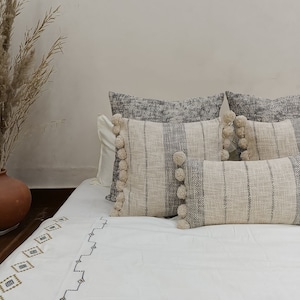 May include: White bed with a white and gray striped throw pillow with pom pom trim. The bed has a white duvet cover with a geometric pattern in brown and black.