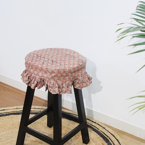 May include: A black wooden stool featuring a round seat cushion with a dusty pink fabric cover. The cover is decorated with a small-scale white floral pattern and finished with a gathered, ruffled skirt that hangs around the edge of the seat.