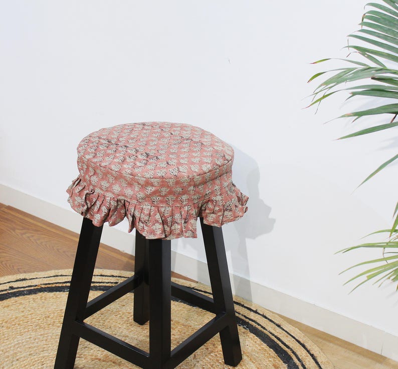 May include: A black wooden stool featuring a round seat cushion with a dusty pink fabric cover. The cover is decorated with a small-scale white floral pattern and finished with a gathered, ruffled skirt that hangs around the edge of the seat.