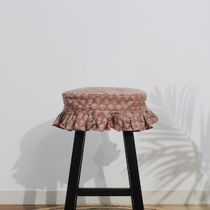 May include: A round stool featuring a black wooden base and a padded seat cushion covered in a dusty pink fabric with a repeating white floral block print pattern and a gathered ruffled skirt edge that hangs around the perimeter of the seat.