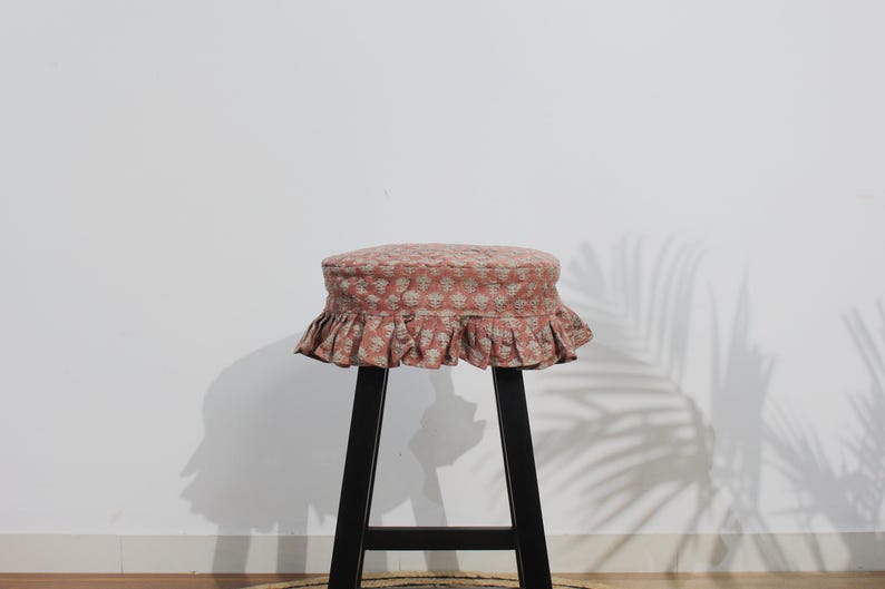 May include: A round stool featuring a black wooden base and a padded seat cushion covered in a dusty pink fabric with a repeating white floral block print pattern and a gathered ruffled skirt edge that hangs around the perimeter of the seat.