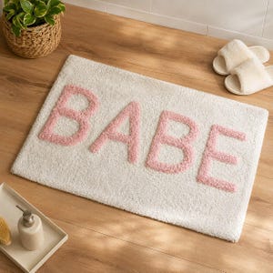 Pode incluir: Um tapete de banho retangular com uma superfície branca texturizada e a palavra BABE escrita em letras grandes, elevadas e cor-de-rosa claro. O tapete é feito de um material macio e felpudo com um design simples e limpo.