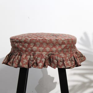 May include: A round stool cover in a muted terracotta red fabric featuring a repeating light gray floral block print pattern. The cover has a quilted top surface and a gathered, ruffled skirt that hangs down around the edges of the stool seat.