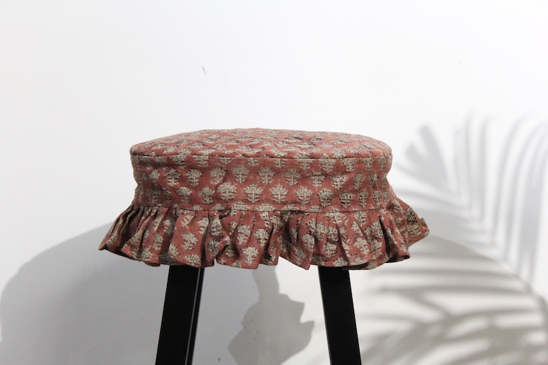 May include: A round stool cover in a muted terracotta red fabric featuring a repeating light gray floral block print pattern. The cover has a quilted top surface and a gathered, ruffled skirt that hangs down around the edges of the stool seat.