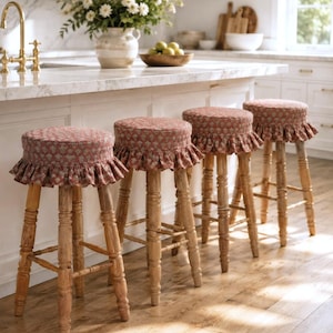 May include: Four wooden bar stools with turned legs and round seats. Each seat is covered in a dusty rose fabric featuring a small-scale repeating floral pattern and a gathered, ruffled skirt that hangs around the edge of the seat cushion.