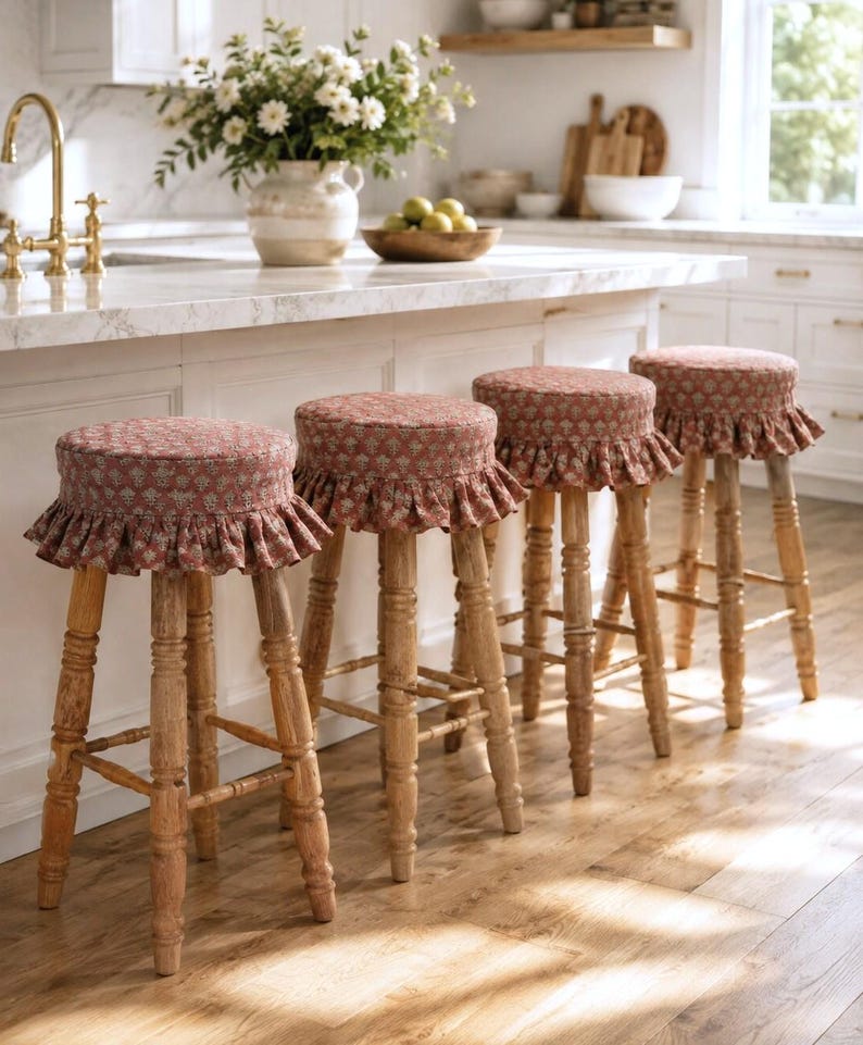May include: Four wooden bar stools with turned legs and round seats. Each seat is covered in a dusty rose fabric featuring a small-scale repeating floral pattern and a gathered, ruffled skirt that hangs around the edge of the seat cushion.
