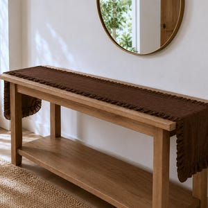 May include: A long, rectangular dark brown table runner with a textured surface and ruffled edges. The runner is draped across the top of a light-toned wooden console table, extending over both ends with a gathered, pleated fabric border.