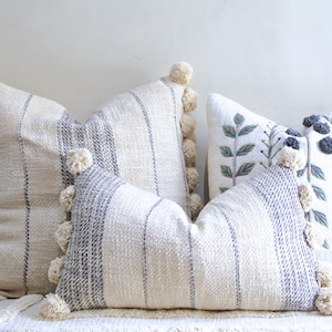 Op de afbeelding: Een verzameling decoratieve sierkussens in verschillende maten en ontwerpen. De kussens hebben crèmekleurige stof met blauwe strepen, geborduurde bloempatronen en crèmekleurige pomponafwerking. De kussens zijn gerangschikt op een wit oppervlak.