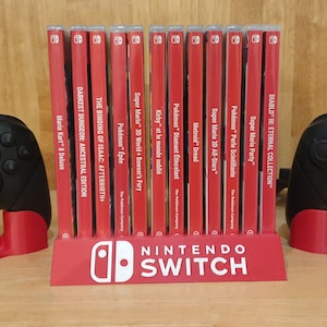 Puede incluir: Un soporte de juegos rojo para Nintendo Switch con un logotipo blanco de Nintendo Switch. El soporte contiene 14 cartuchos de juegos de video, incluidos títulos como "Super Mario Party", "Pokémon Espada" y "Metroid Dread". Dos mandos negros de Nintendo Switch están a cada lado del soporte.