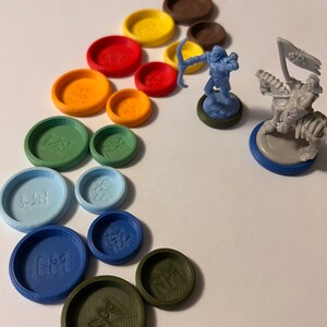 Puede incluir: Un conjunto de fichas de juego de pl&aacute;stico de colores con diferentes s&iacute;mbolos, una figura en miniatura azul de un arquero y una figura en miniatura gris de una criatura montando un caballo. Las fichas est&aacute;n dispuestas en forma de arco&iacute;ris.