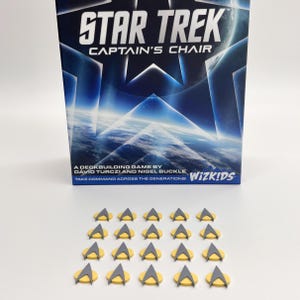 以下が含まれることがあります： 「STAR TREK CAPTAIN'S CHAIR」のテキストが入った、Star Trek Captain's Chairのデッキ構築ゲームボックス。下には、黄色とグレーのスターフリート司令部記章トークンがいくつかあります。ゲームはDavid TurcziとNigel Buckleによるものです。