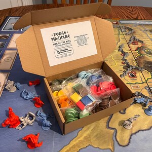 Puede incluir: Una caja de cart&oacute;n llena de marcadores de base en miniatura para el juego "War of the Ring" de Forge Machine. La caja est&aacute; abierta y los marcadores son visibles en bolsas de pl&aacute;stico. Los marcadores son de varios colores, incluyendo rojo, azul, verde, amarillo y marr&oacute;n.