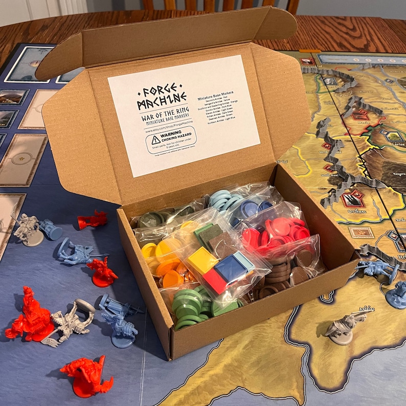 Puede incluir: Una caja de cart&oacute;n llena de marcadores de base en miniatura para el juego "War of the Ring" de Forge Machine. La caja est&aacute; abierta y los marcadores son visibles en bolsas de pl&aacute;stico. Los marcadores son de varios colores, incluyendo rojo, azul, verde, amarillo y marr&oacute;n.