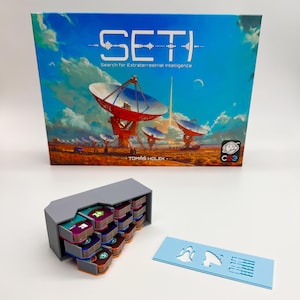 Puede incluir: Una caja gris con un juego de cartas para el juego de mesa "SETI: Search for Extraterrestrial Intelligence". La caja está abierta y las cartas son visibles. La caja del juego está en el fondo con una ilustración colorida de un radiotelescopio.