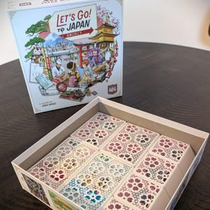 May include: A board game box titled "Let's Go! To Japan" with colorful illustrations of Japanese landmarks and people. The open box reveals game components with floral cutouts. The game is by Josh Wood.