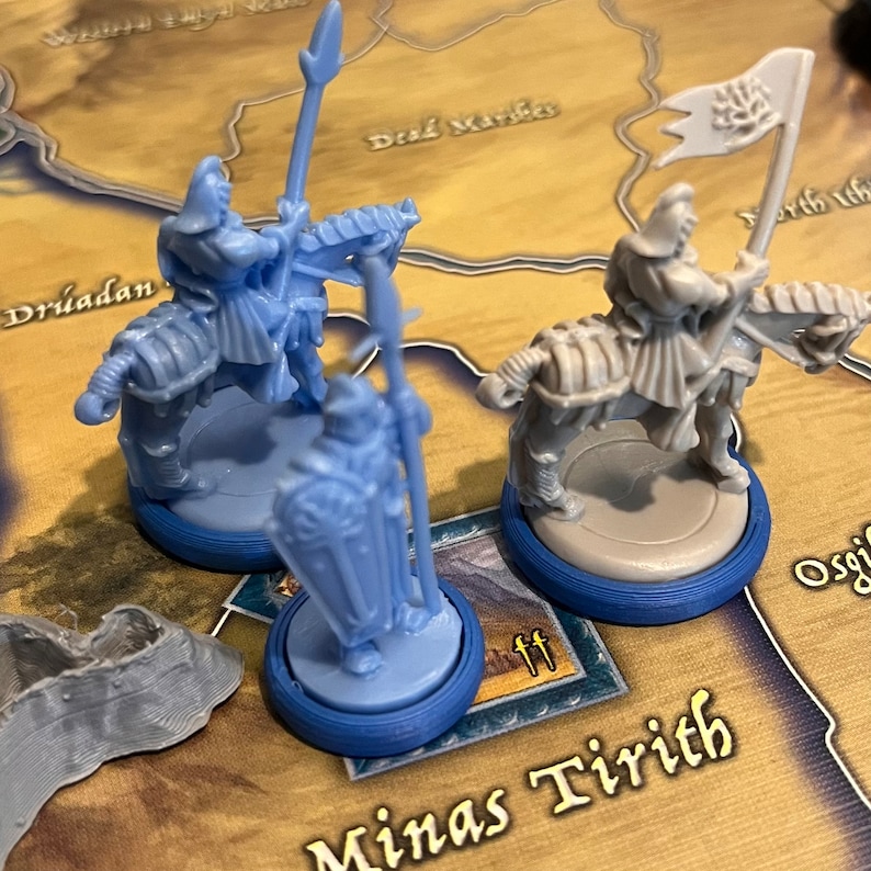 Puede incluir: Tres figuras en miniatura de pl&aacute;stico de caballeros montados, dos son azules y uno es gris. Las figuras est&aacute;n sobre bases circulares con anillos azules y grises. Las bases est&aacute;n sobre una superficie marr&oacute;n con un mapa y las palabras "Minas Tirith" impresas en &eacute;l.